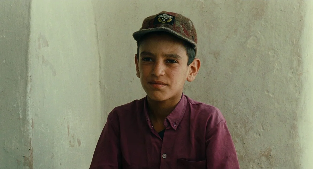 The Wind Will Carry Us (1999) movie still: medium — The subject stands in front of a plain, light-colored textured wall. A young boy wearing a cap…