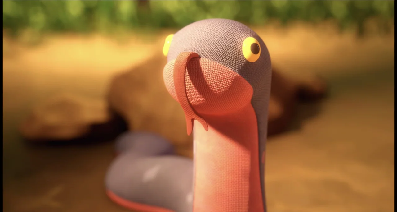 The Wishmas Tree (2020) movie still: closeup — A fabric-textured snake puppet rests on a sandy surface near green plants. The puppet maintains an…