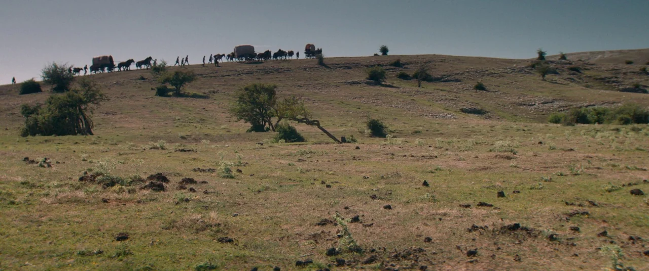 The Sisters Brothers (2018) movie still: wide — A vast, open prairie stretches beneath a clear sky with rolling hills in distance. A small line of…