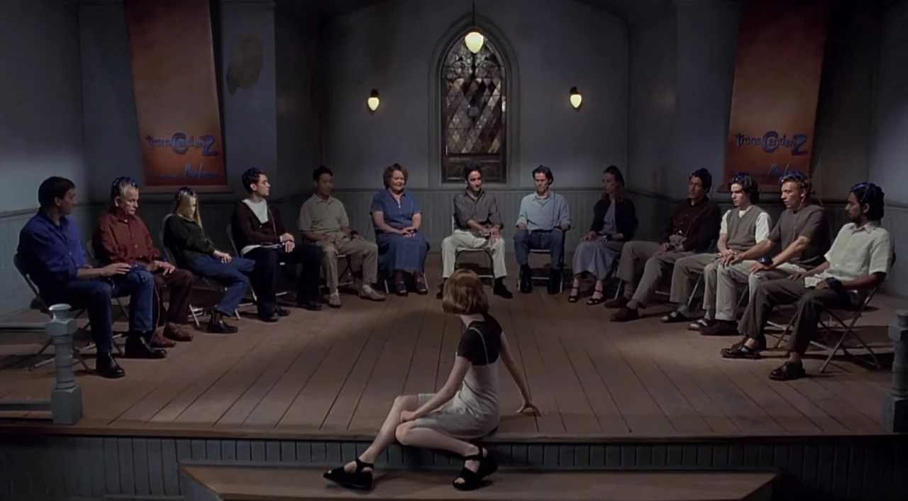 eXistenZ (1999) movie still: wide — A community center meeting room features a wooden stage and rows of folding chairs. A young woman sits alone on…