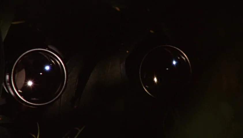 A Bay of Blood (1971) movie still: closeup — A dark, undifferentiated background frames two circular optical lenses. Light reflects off the glass…