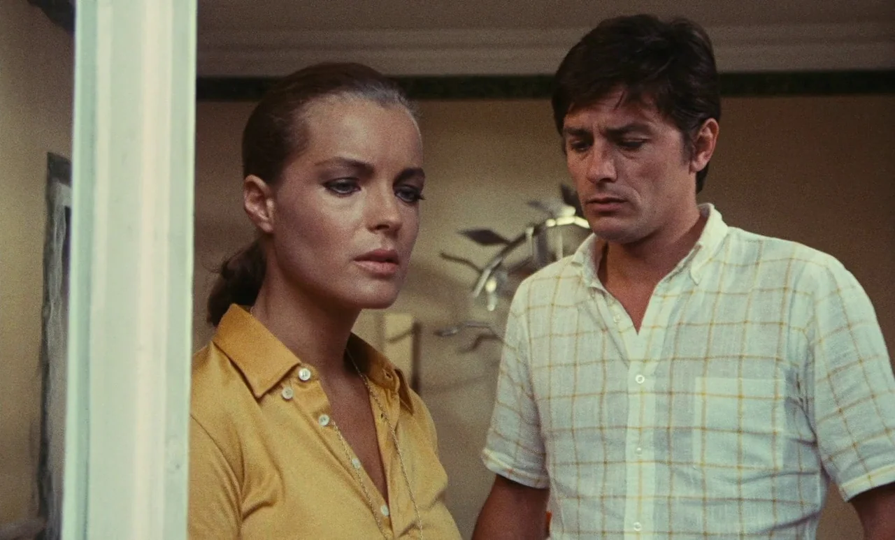 The Swimming Pool (1969) movie still: medium — An interior space features a woman in yellow and a man in a grid-pattern shirt. They stand close…