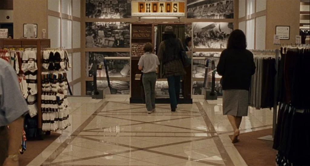 Choke (2008) movie still: fullbody — A 1990s retail corridor features beige tiled flooring and racks of hanging clothing. Three people walk away…