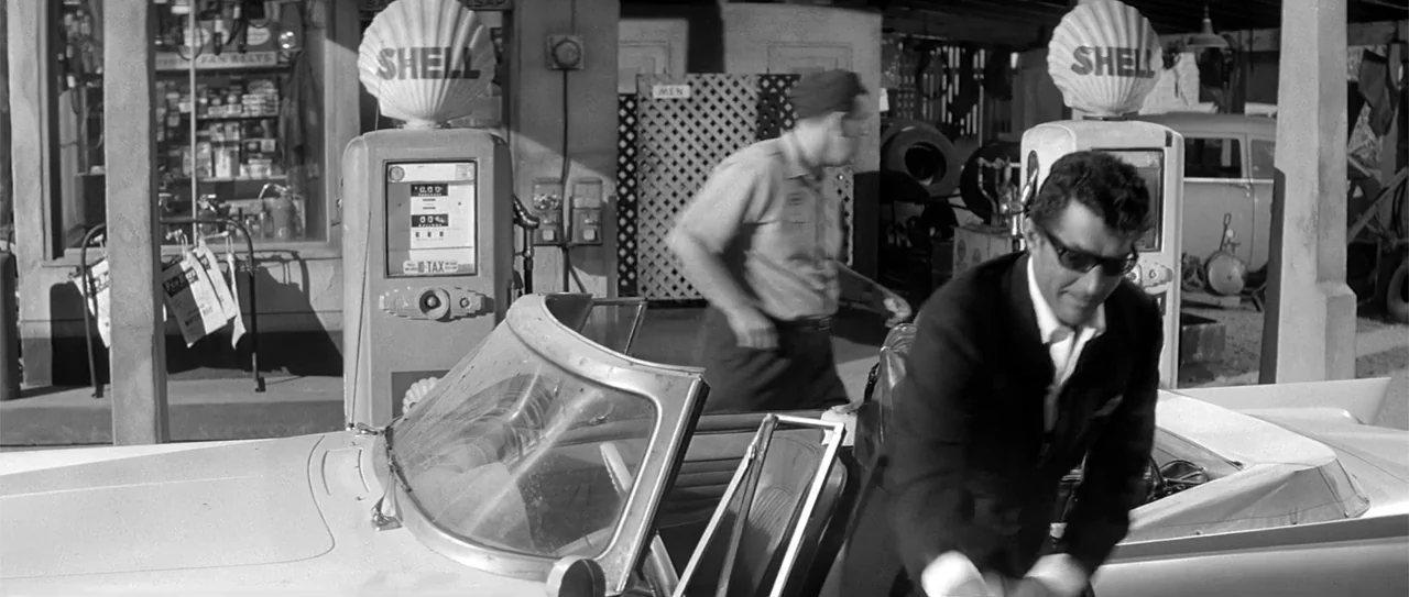 Kiss Me, Stupid (1964) movie still: medium — A 1950s service station features Shell branded pumps and a parked convertible car. A man in a suit…