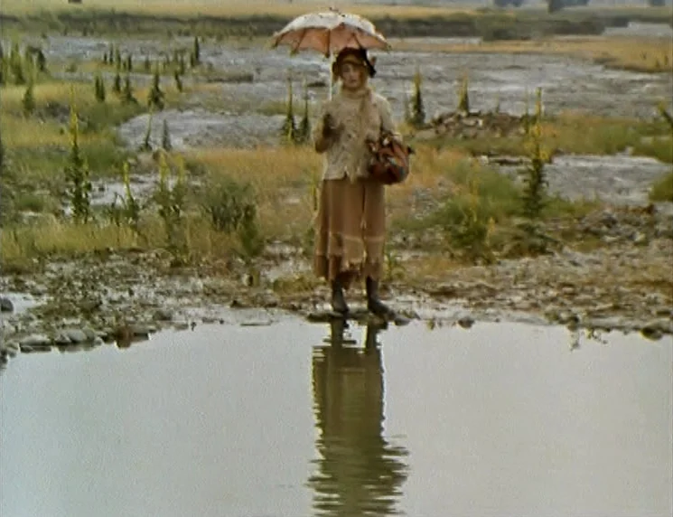 The Wishing Tree (1977) movie still: fullbody — A sparsely vegetated, rocky riverbank with patchy grasses lies under an overcast, hazy sky. A woman…