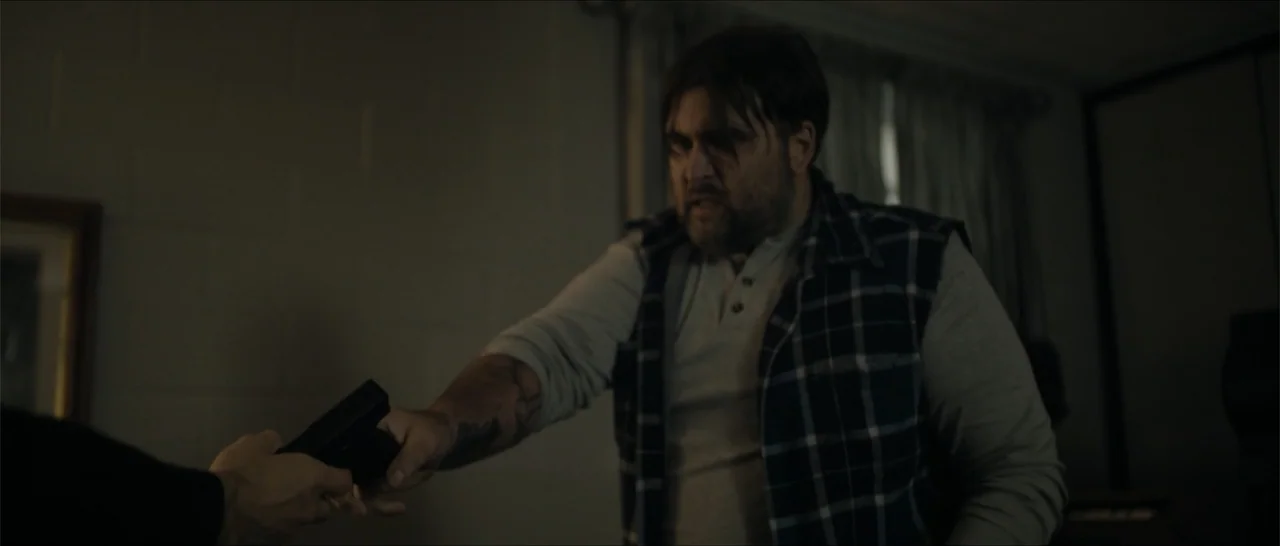 Crescent City (2024) movie still: medium — A dimly lit room with plain walls and a drawn curtain in the background. A man points a handgun toward an…