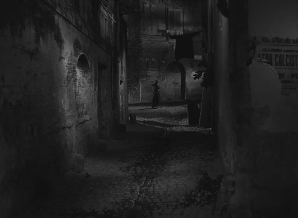 Il Bidone (1955) movie still: wide — A dark cobblestone narrow path lies between weathered stone buildings in a European town. A single person…