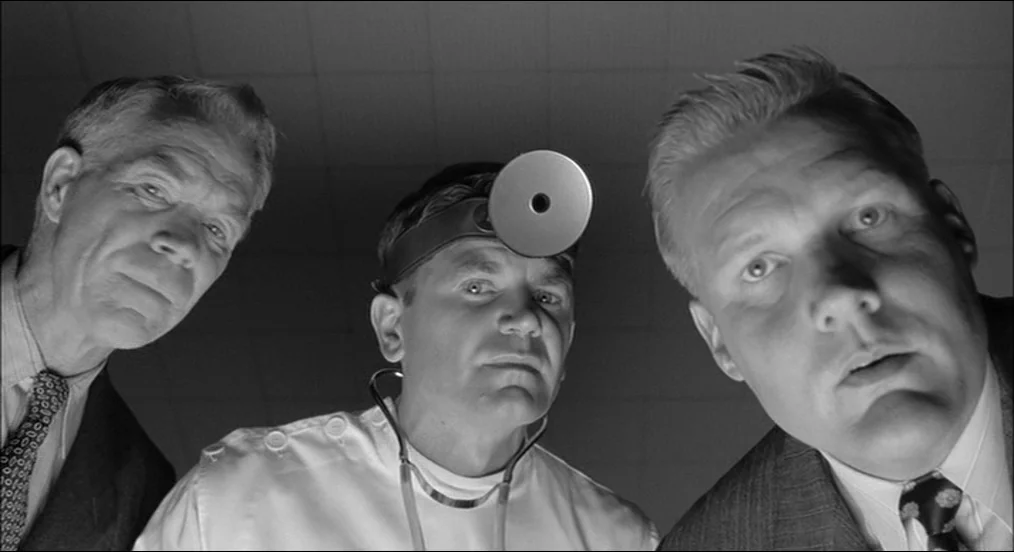 The Man Who Wasn’t There (2001) movie still: medium — Three men stand over the camera in a sterile white-tiled medical space. The central figure…