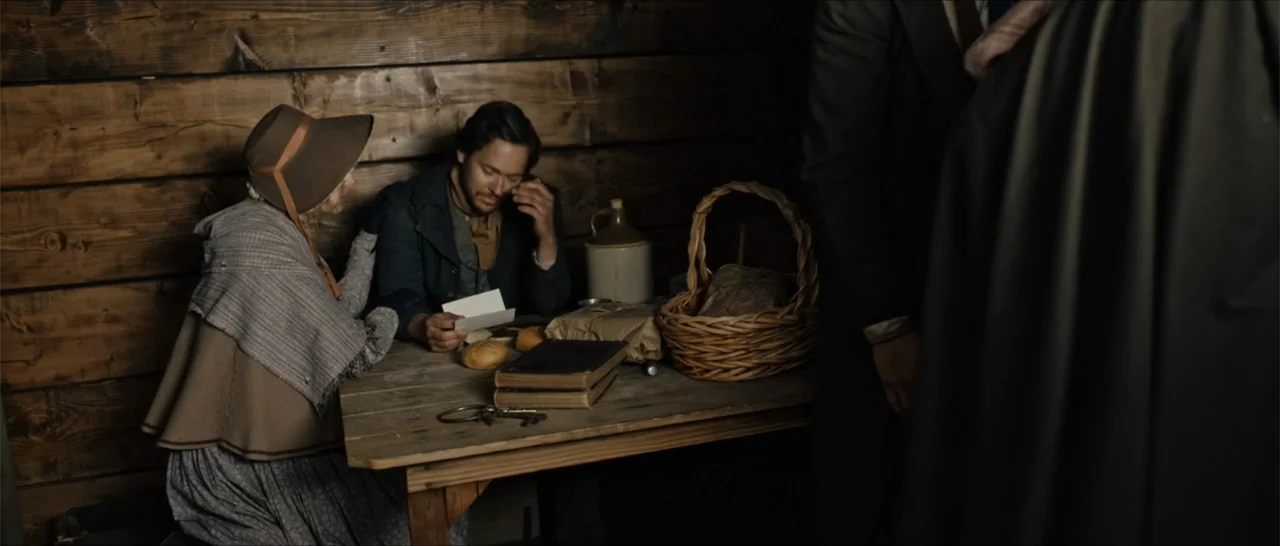 Out of Liberty (2019) movie still: medium — A dimly lit log cabin features dark wood walls and a wooden table. A man and woman sit at the table with…