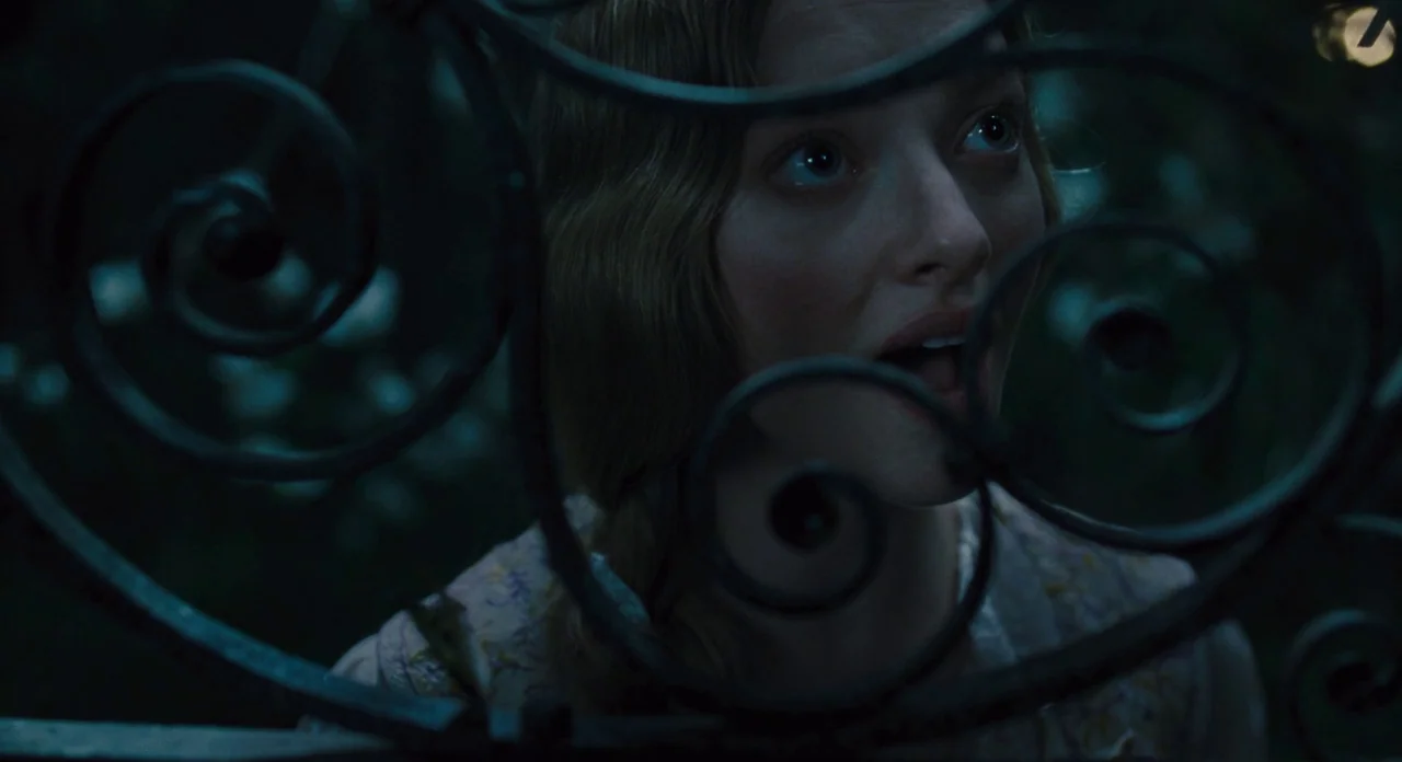 Les Misérables (2012) movie still: closeup — A dark outdoor space is framed by the intricate scrolls of a heavy iron gate. A young woman with light…