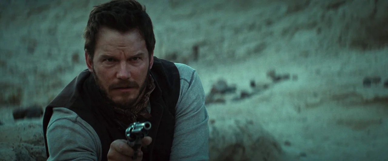 The Magnificent Seven (2016) (2016) movie still: medium — A man crouches in a sandy, desolate desert terrain with exposed dirt textures. He aims a…