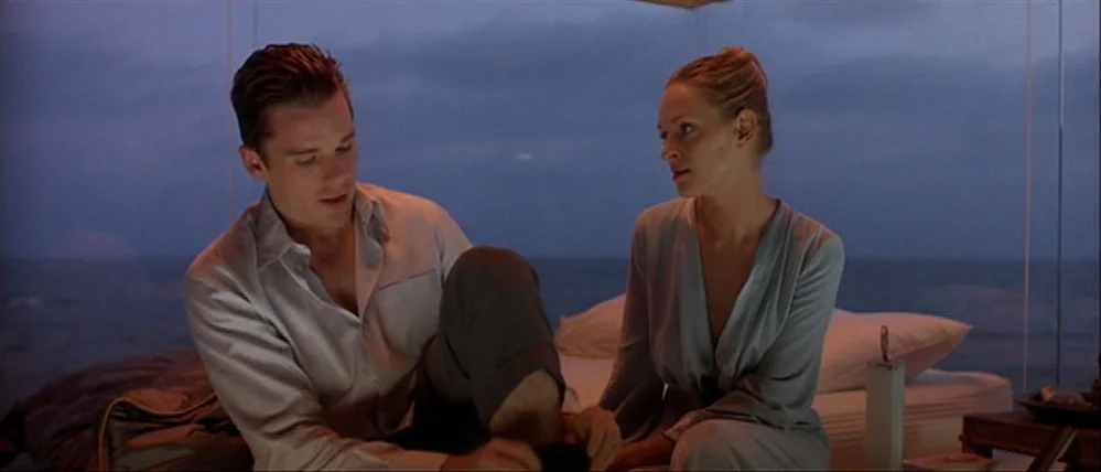Gattaca (1997) movie still: medium — A man and woman sit on a bed within a glass-walled room at dusk. The ocean is visible directly outside the…