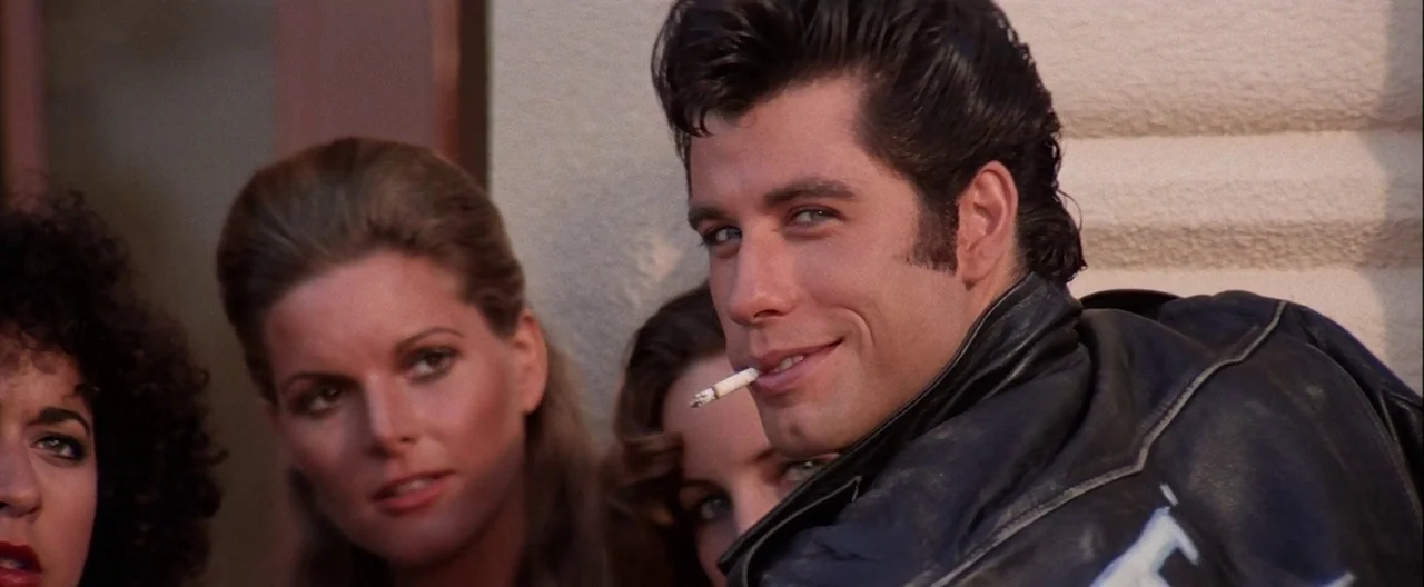 Grease (1978) movie still: medium — A group of people stands before a light-colored stucco wall in daylight. A young man in a leather jacket holds a…