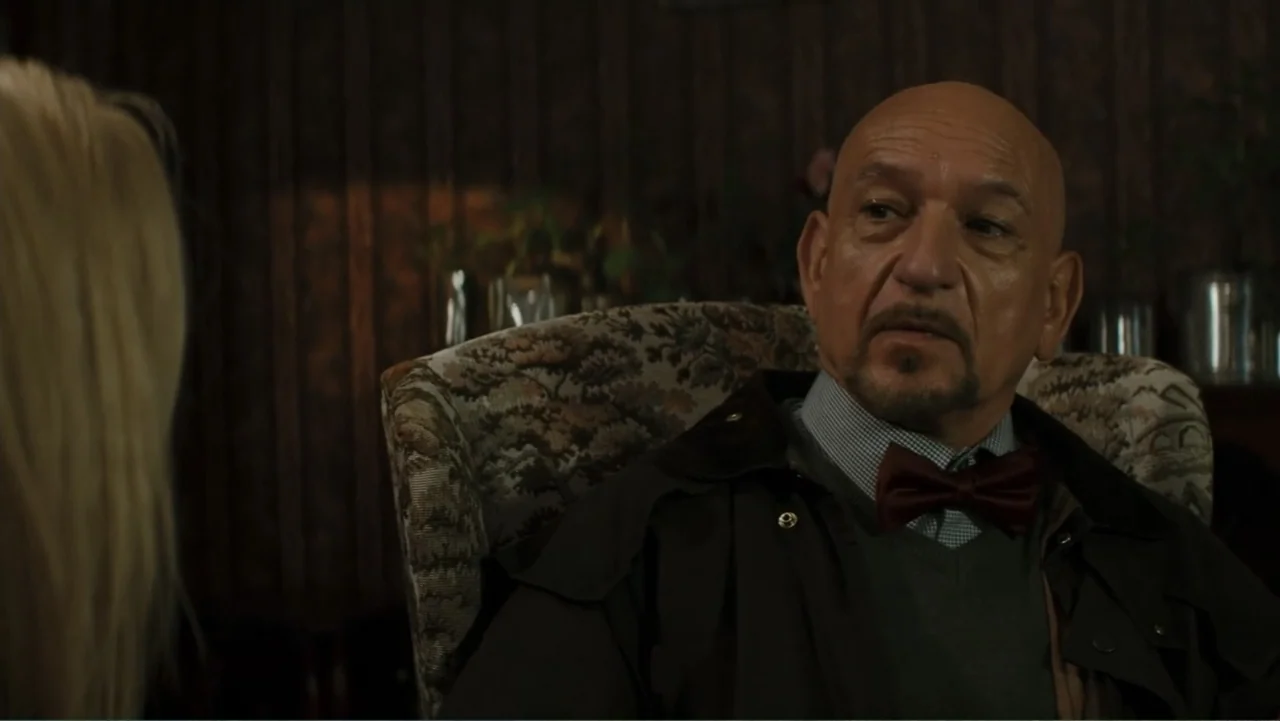 Robot Overlords (2015) movie still: medium — A dimly lit room features a man sitting in a vintage fabric armchair. The man wears a brown jacket and…