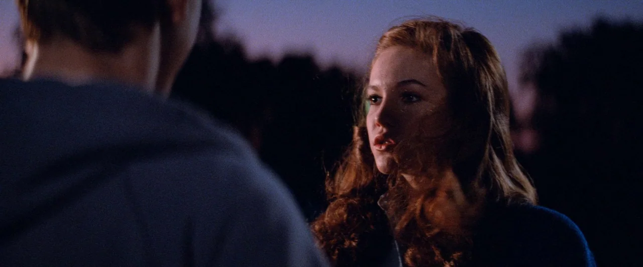 The Outsiders (1983) movie still: medium — A dark outdoor space at dusk features blurred silhouettes of trees in the background. A young woman with…