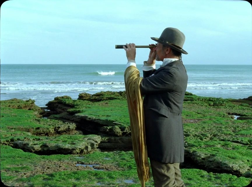 Jauja (2014) movie still: medium — A man stands on a moss-covered rocky shoreline next to the ocean waves. He holds a brass spyglass to his eye…