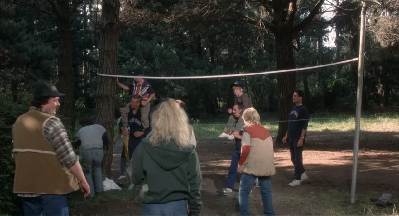 Overboard (1987) movie still: wide — A wooded area with tall pine trees and a thin white net stretched across. Several people in casual 1980s attire…