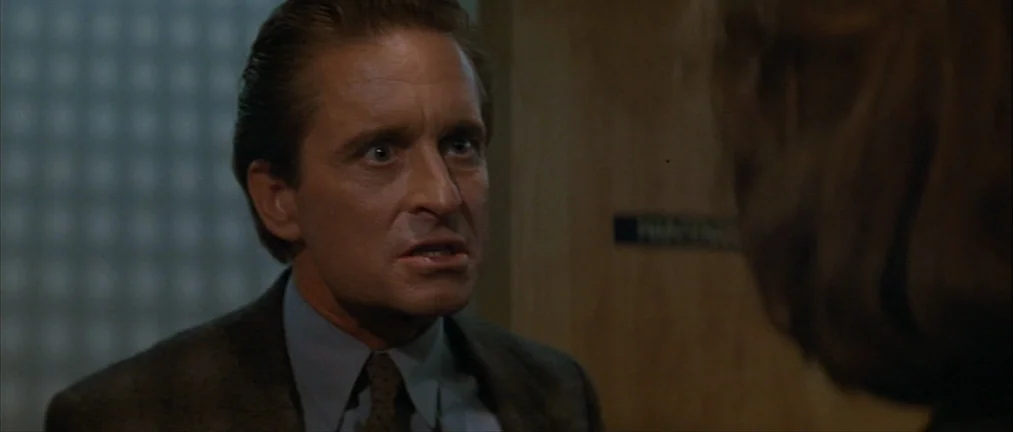 Basic Instinct (1992) movie still: closeup — A dark office environment features wooden wall paneling and a small door sign. A man in a suit looks…