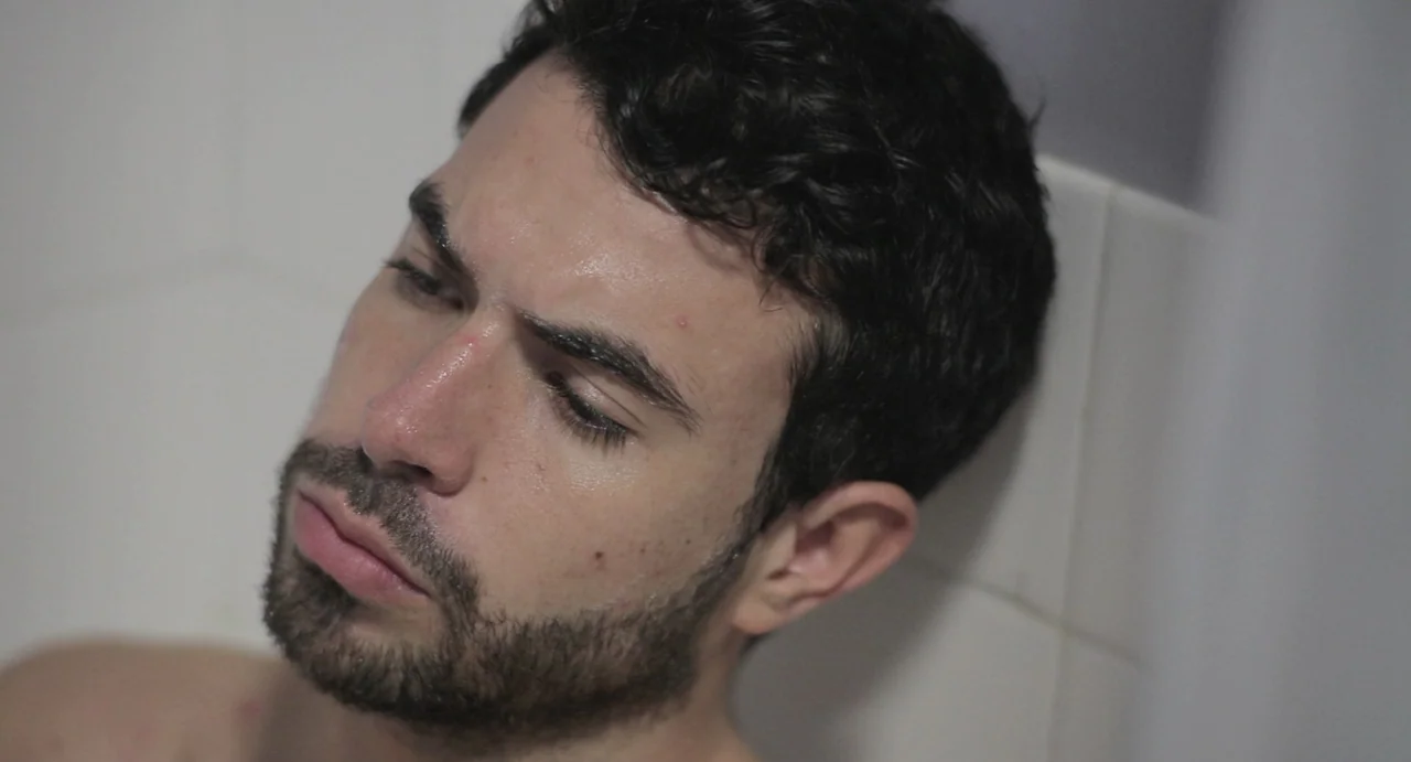 Weekend (2011) movie still: closeup — A close-up of a young man lying in a white-tiled bathtub. The man has his eyes closed and his head is resting…