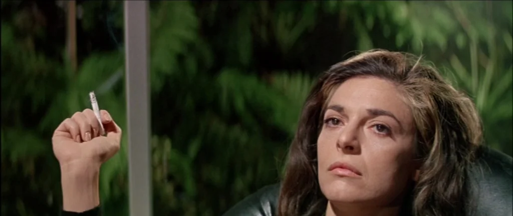 The Graduate (1967) movie still: closeup — A woman sits in a dark room with blurred green foliage visible behind her. She holds a cigarette in her…