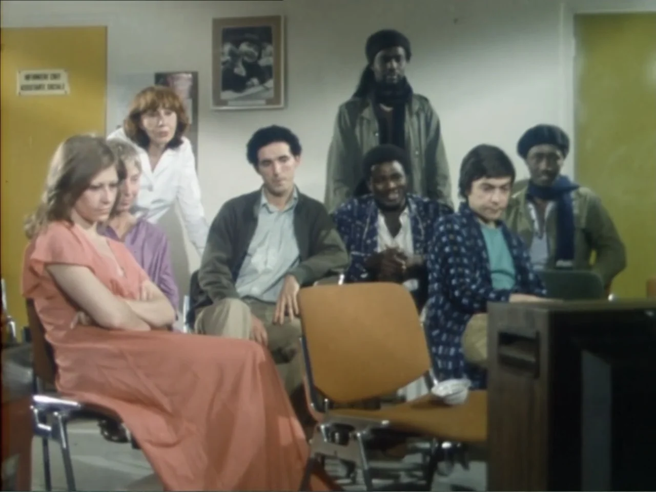 A Dessert for Constance (1981) movie still: medium — A group of people sit on chairs in a brightly lit, sterile room. Individuals wait together in a…