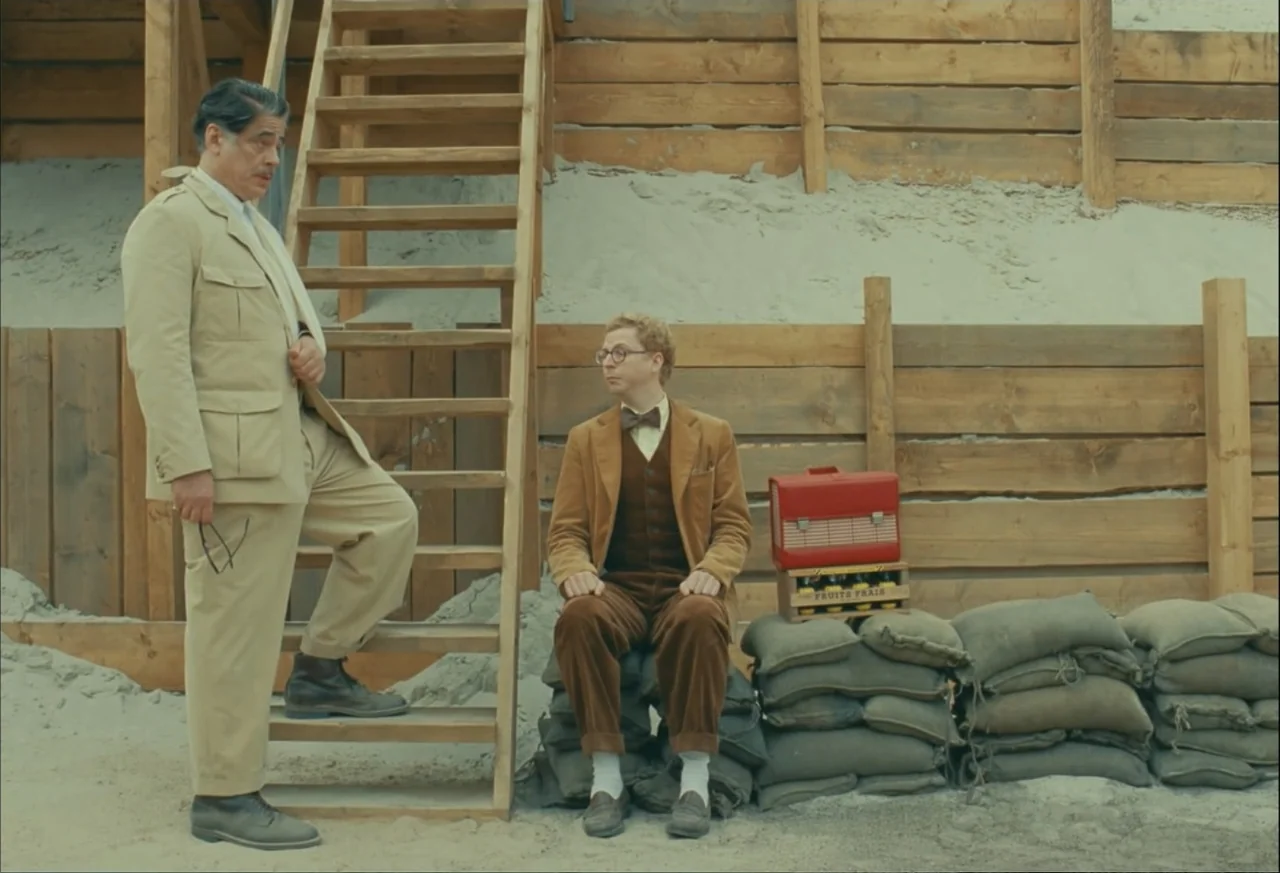 The Phoenician Scheme (2025) movie still: fullbody — A sandy construction area features a wooden retaining wall and a set of stairs. Two men…