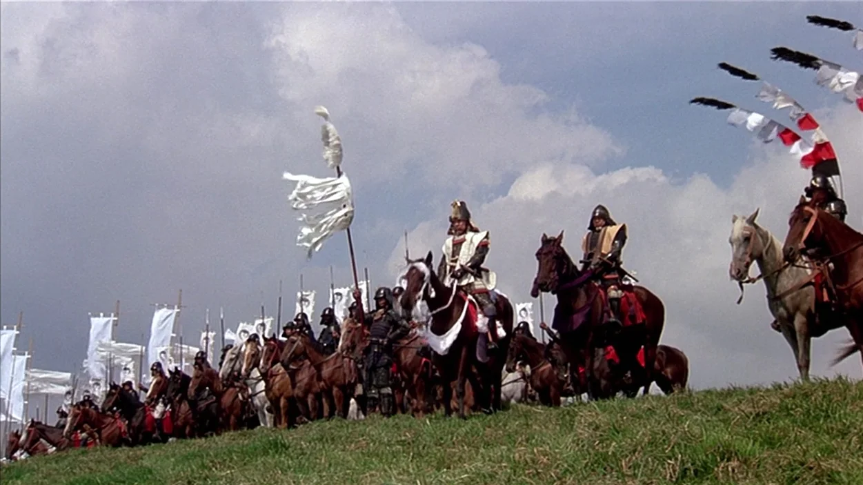 Ran (1985) movie still: wide — A line of mounted samurai warriors in traditional armor stands upon a grassy hill. Numerous white flags and colorful…