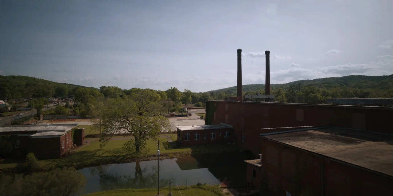 The Pickup (2025) movie still: establishing — An industrial complex features brick buildings with two tall smokestacks and a reflecting pond. A…