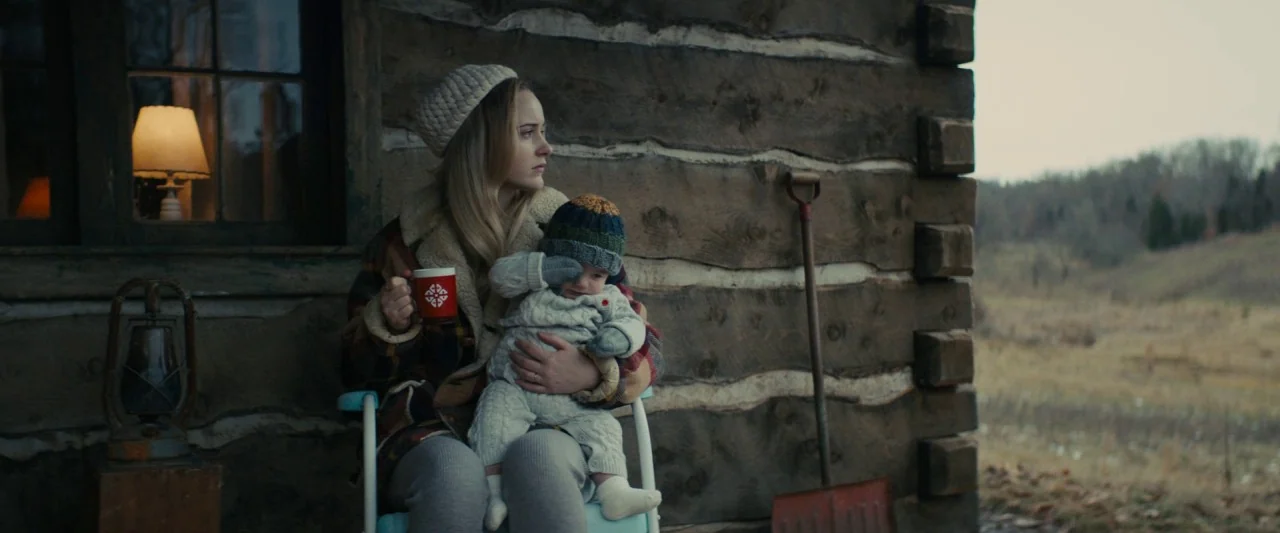 I'm Your Woman (2020) movie still: medium — A woman and baby sit on a metal chair against a rustic log structure wall. A red mug and a shovel rest…