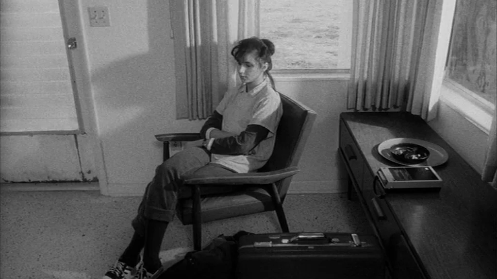 Stranger Than Paradise (1984) movie still: medium — A spartan motel room with simple furniture including a wooden chair and side table. A woman sits…