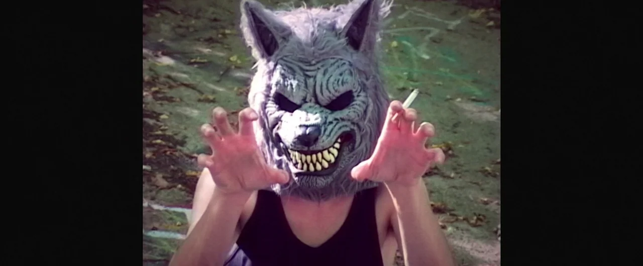 Poser (2021) movie still: closeup — A person wears a textured gray wolf mask while positioned in an outdoor space. The subject holds their hands up…