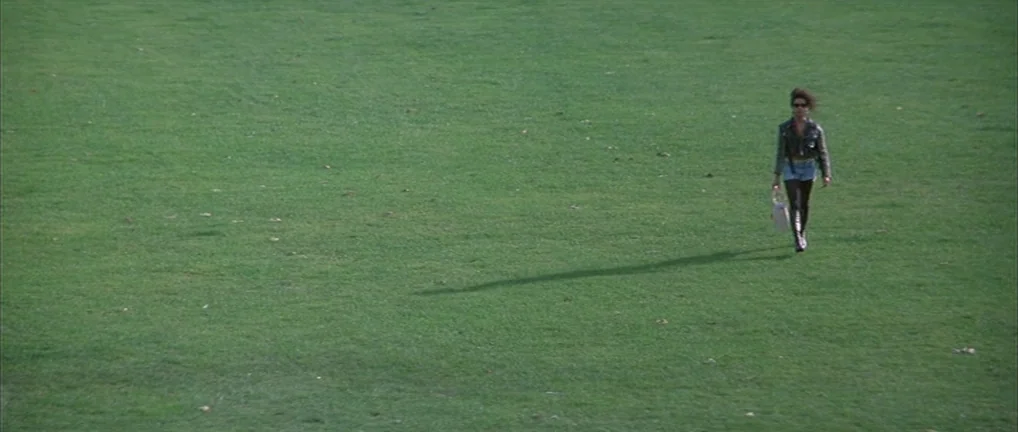 The Crying Game (1992) movie still: wide — A vast expanse of flat, green mown grass covers the entire frame. A lone woman walks across the field…