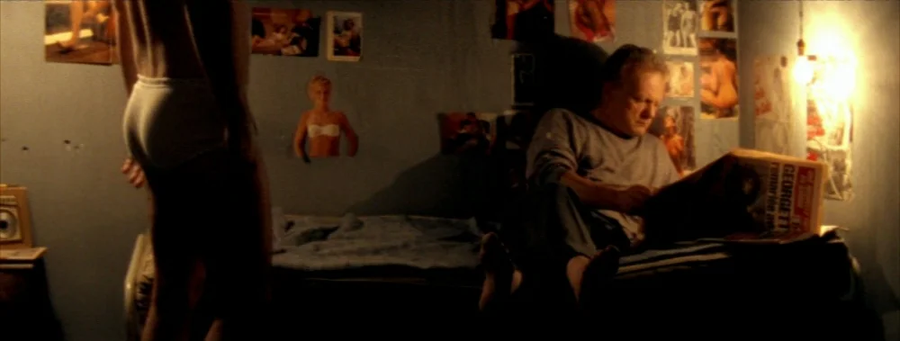 Carne (1991) movie still: medium — A dark, dimly lit room with walls covered in various pinned photographs and posters. A man sits on a bed reading…