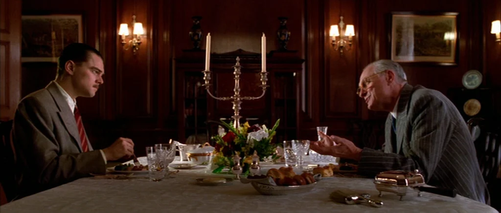 The Aviator (2004) movie still: medium — A dimly lit room features wood-paneled walls and a large, formal dining table. Two men sit facing one…
