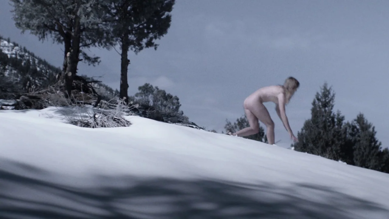Snowbound (2017) movie still: wide — A desolate snow-covered hillside features a few scattered evergreen trees under a grey sky. A nude woman moves…