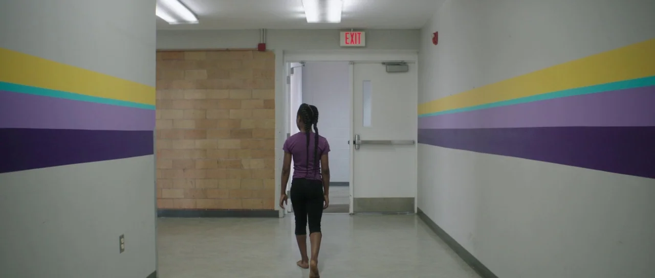 The Fits (2016) movie still: fullbody — A sterile school hallway features walls with painted horizontal bands of yellow, teal, and purple. A young…