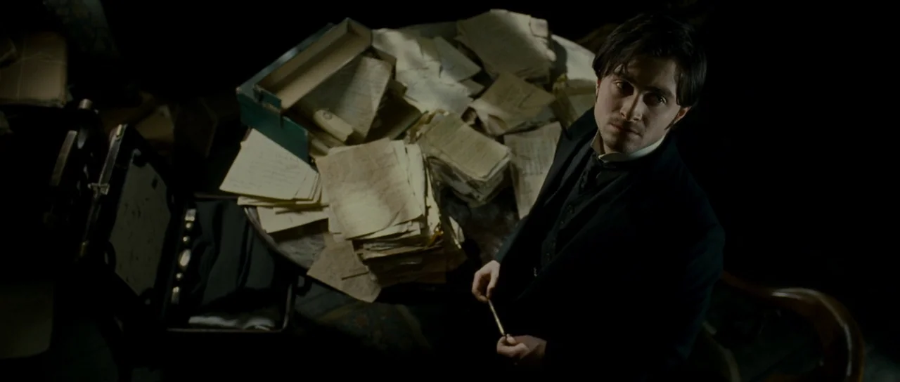 The Woman in Black (2012) movie still: medium — A dark, dimly lit study is filled with stacks of old paper documents. A young man stands beside the…
