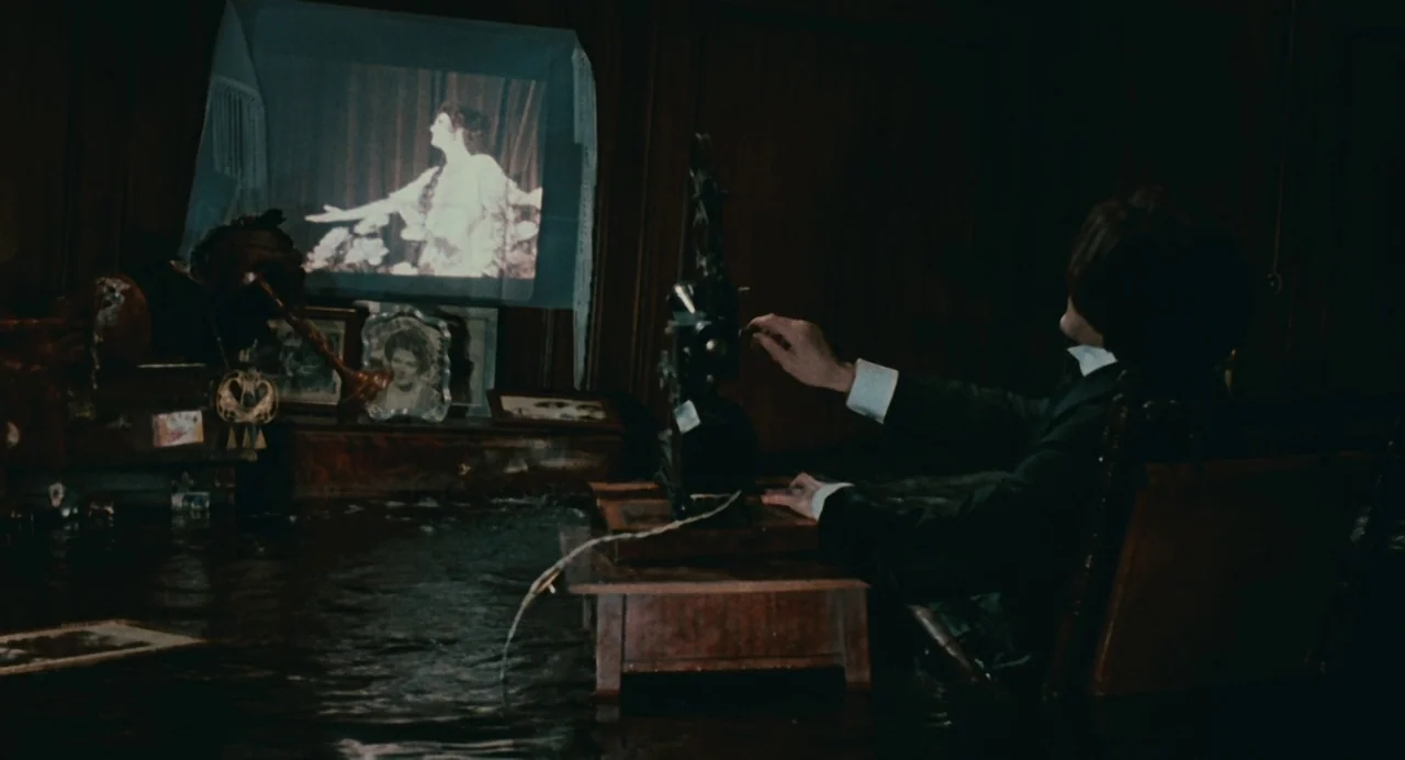 And The Ship Sails On (1983) movie still: medium — A dark flooded room contains a man seated at a wooden desk with a projector. The projector…