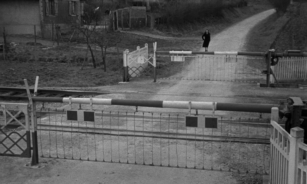 Léon Morin, Priest (1961) movie still: wide — A rural level railroad crossing with striped wooden gates lowered across the gravel road. A woman…