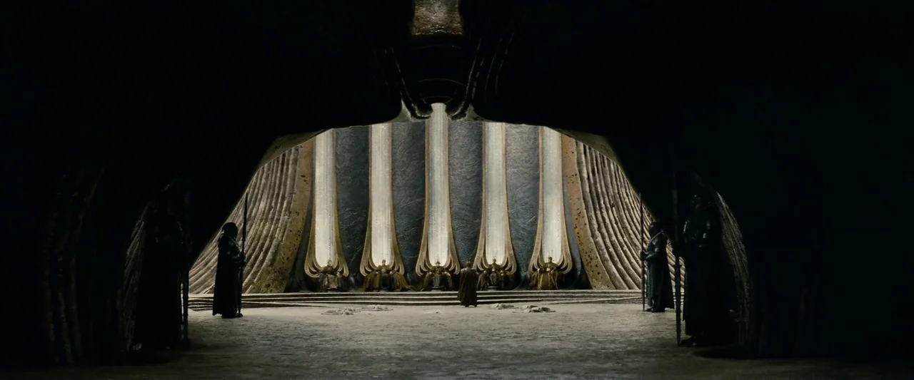 Man Of Steel (2013) movie still: wide — A dark stone chamber features five throne-like niches backed by vertical illuminated light panels. A small…