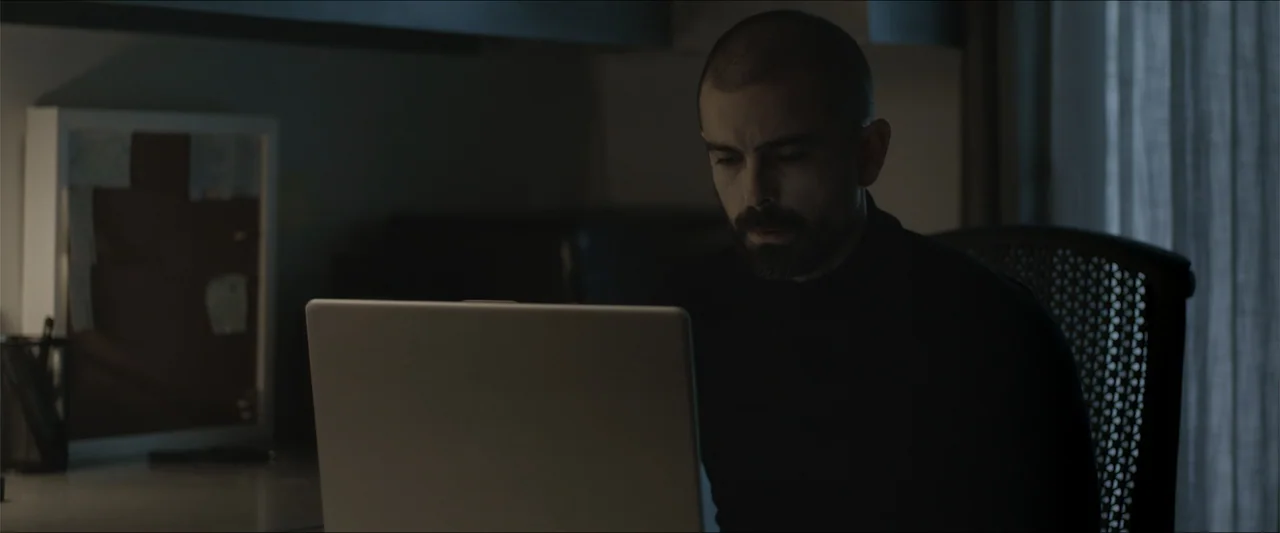 Los habitantes (2023) movie still: medium — A dimly lit home office with a dark desk and a curtained window visible. A man with a short-cropped…