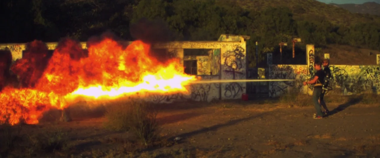 Bellflower (2011) movie still: wide — A graffiti-covered, crumbling concrete structure sits in a dry, sparse clearing at sunset. A man fires a large…