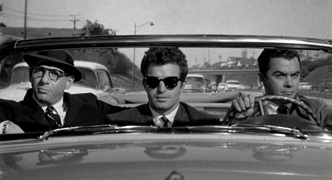 Murder By Contract (1958) movie still: medium — Three men in suits sit in the front row of an open-top vehicle. The setting is a bright, sunny…