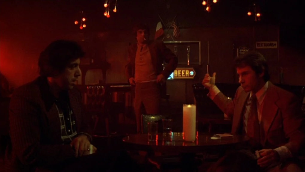 Mean Streets (1973) movie still: medium — A dimly lit 1970s bar with wood-paneled walls and a neon beer sign. Two men sit at a round table having a…