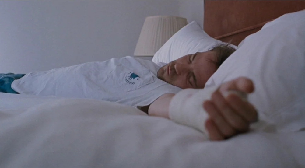 Somewhere (2010) movie still: medium — A man sleeps on a white bed with pillows and a reddish-brown headboard. He has a white cast on his arm while…