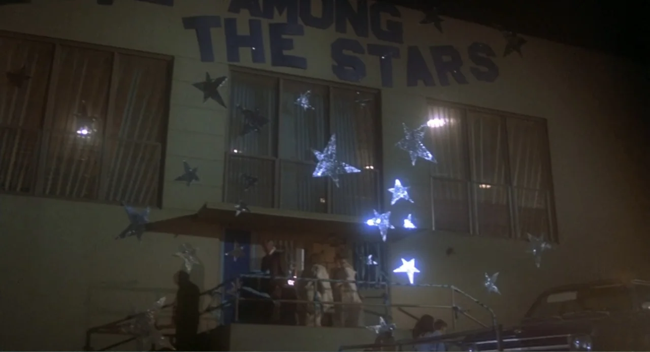 Carrie (1976) movie still: wide — A two-story building features the text 'AMONG THE STARS' painted above large glass windows. Several glowing white…