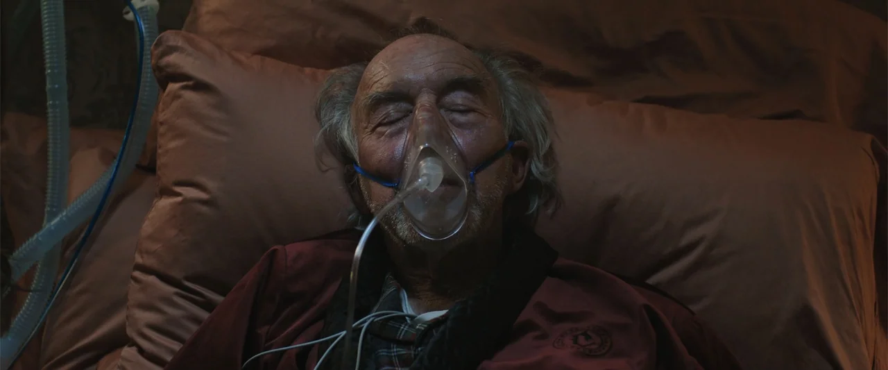 Trap House (2025) movie still: closeup — A dimly lit bedroom features an elderly man resting on brown bedding. The man wears an oxygen mask while…