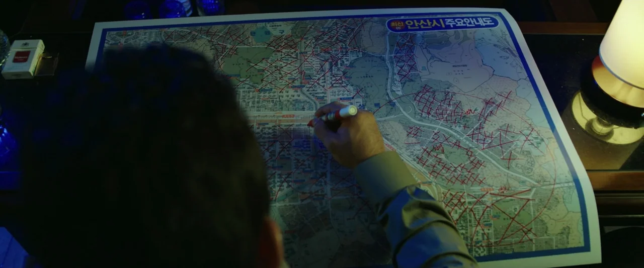 The Gangster, the Cop, the Devil (2019) movie still: medium — An interior space features a large paper city map laid flat on a dark surface. A…