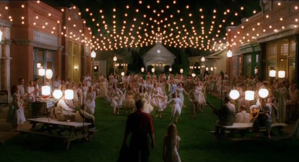 Big Fish (2003) movie still: wide — A town square at night features a large gathering of people dancing on grass. Overhead strings of lights…