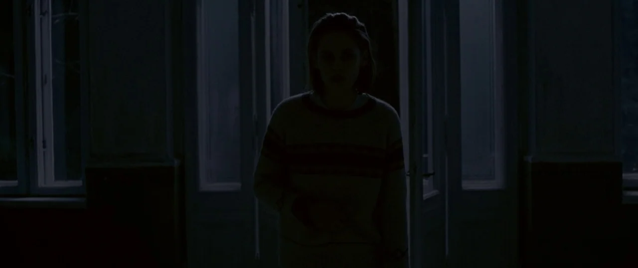 Personal Shopper (2016) movie still: medium — A woman stands centered in a dark room with windows visible in the background. She is illuminated only…
